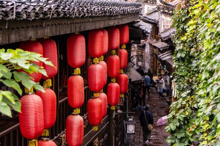 Red Chinese lanterns made from paper with gold tassels are being hang from the roof of the old Chinese styled building and many tourists walking with umbrella through the alley full with shops on the rainy day located at the street in Fenghuang Province in Chinaの写真素材