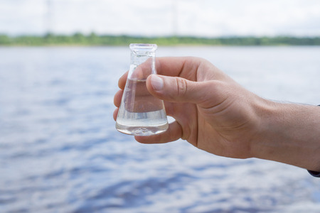 Water Purity Test. Hand holding a chemical flask with water, lake or river in the background.の写真素材