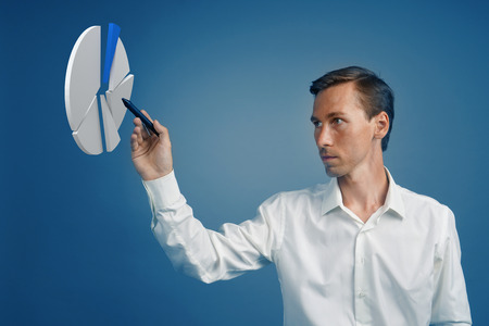 Young man in white shirt working with pie chart on blue background. Concept on the topic of statistics or financial report.の写真素材