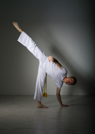 Young Man in white sportswear performing a kick. Indoor martial arts training, capoeira.の写真素材