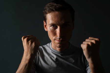 Young man in t-shirt on a dark background holding his fists at his face. Photo in low key. Low Key...