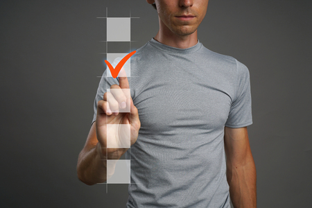Young man in t-shirt checking on checklist box. Gray background. Concept on the topic of elections.の写真素材