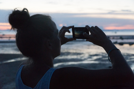 Woman takes a sunset above the sea on a phone camera.の写真素材