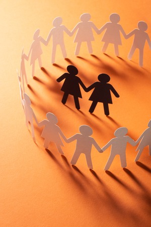 Paper figure of female couple surrounded by circle of paper people holding hands on red surface....