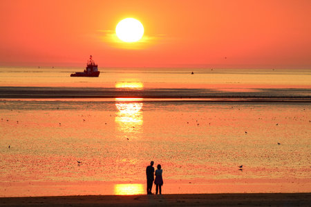 A beautiful and rare sunset at the beach during the summer heatwave. A couple in love are gazing at the falling sun as a boat approaches. The sun reflection can be seen in the seaの写真素材