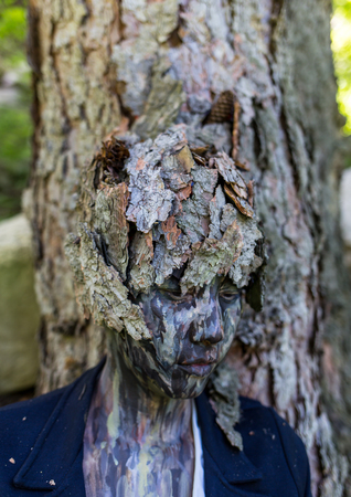 Girl Office worker in a business suit, outdoors with a makeup on her face imitating the bark of a tree. The girl merges with the tree trunk, sits and rests her back on him.の写真素材