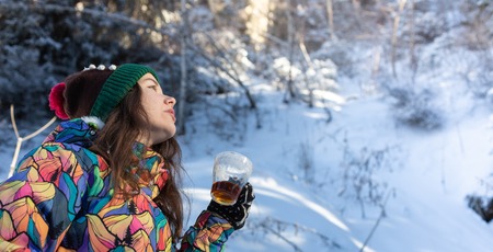 Girl enjoys the snow falls. Young woman in a knitted shape is drinking tea in the forest during a snowfall. Toned photoの写真素材