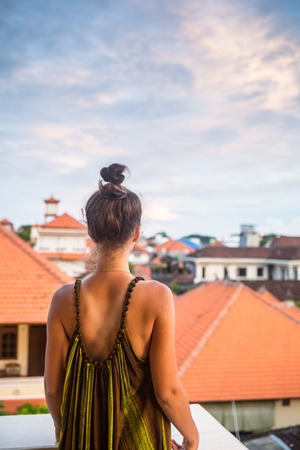 Beautiful young woman sitting on a mountain, back view. Below - the sea and the tiled roofsの写真素材