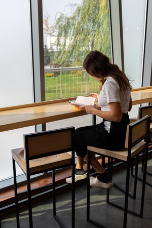 Positive young woman in stylish eyeglasses dreaming while sitting at table with opened book in...