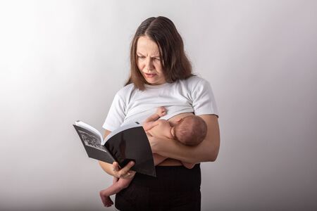 Beautiful young mother breastfeeding a baby while reading a black bookの写真素材