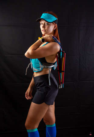Beautiful girl marathon runner in blue jogging equipment and trekking poles in the studio on a black backgroundの写真素材