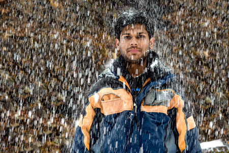 Young, Fair and Handsome Indian boy in his early 20 in skiing costume, wearing snowsuit, standing still and posing in neutral mood during heavy snowfall in winters during the onset of Christmasの写真素材