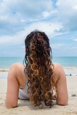 Amazing Back view of fair skinned Girl in mid 20s, having curly hairs of golden Color. She is relaxing & sunbathing solo on beach vacation, to exotic tropical island with clement weather & calm ocean.の写真素材