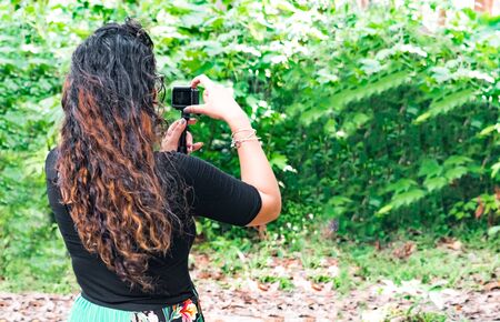 Trendy brunette botanist in her 20s, with curly hairs, is monitoring natural growth of plants during outdoor field work, and is exploring the natural beauty while clicking picture from action camera.の写真素材