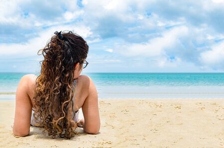 Back view pose of attractive and beautiful young girl in her early 20s with long and colored curly hairs, while getting tanned at beach on a bright sunny day, during her holidays on tropical island.の写真素材