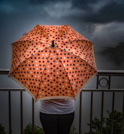 Amazing photo of rainy season of monsoons, wherein an unrecognizable teenage girl is holding a peach colored umbrella with polka dots design, while standing on a  cliff edge, enjoying the clouds.の写真素材