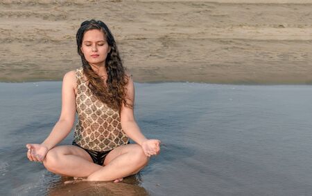 Gorgeous young girl in her early 20s, is meditating for her physical and mental well being, while sitting in lotus position on beach. Concept of health, yoga, well being, lifestyle, relaxation etc..の写真素材