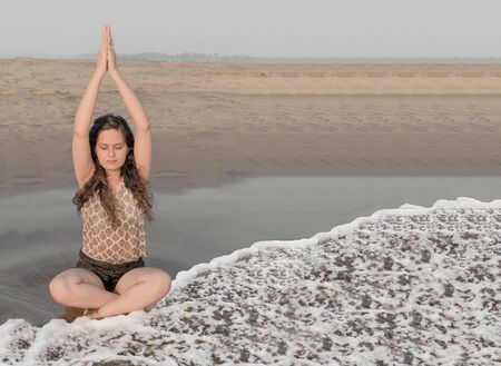 Beautiful photo of pretty young girl practicing yoga on beach in morning hours by raising both her hands up in prayer pose to relax her body, mind and soul while sitting in padmasana or lotus pose.の写真素材