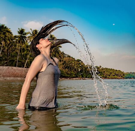 Perfectly timed shot of Caucasian Girl with black hairs in her early 20s, wearing sleeveless Spaghetti Top and enjoying her solo trip at island, by playing in sea and whipping her wet hair off waterの写真素材
