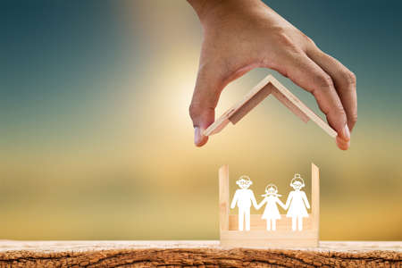 Mah hand hold to open a roof and wooden home with happy family of paper art is placed inside on nature sunlight, The saving money for house or business real estate owner in the future concept.の写真素材
