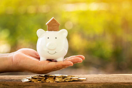 Investor hand hold a piggy bank with the home model put on the top and coin put on the old wood in the public park, Loan or saving money  for buy a new house or investment the real estate concept.の写真素材