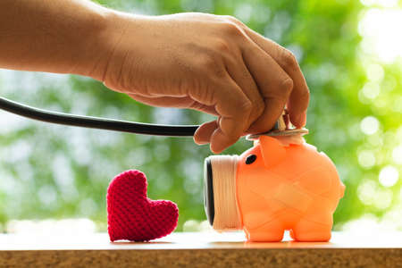 Man hand hold is use a stethoscope with check hygiene of a sick piggy bank and a  red heart put on...