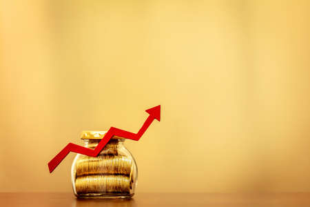 Bottle bank gold coin and red arrow graph with growing value put on the desk on brown background,...