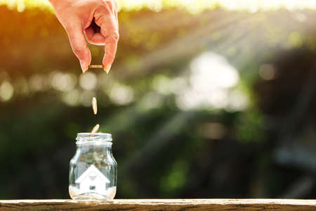 Investor hand hold and drop a gold coin in the bottle and home model with savings money for growing in the public park, Business investment for real estate and or loan for buy house concept.の写真素材