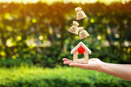 Woman hand holding a home model tied with red ribbon and money bag drop on the top in the public park, The buying a new real estate as a gift to family or the one loved concept.の写真素材