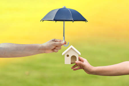 Woman and man hand hold the wooden home model and open the black umbrella for protect on sunlight in the public park, Saving money for real estate and property protection concept.の写真素材