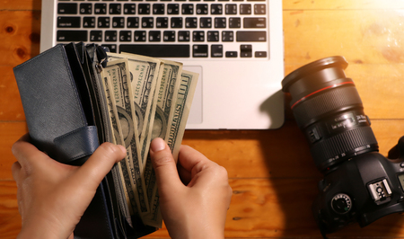 Desktop flatlay: laptop, camera, womanâs hand holding wallet with one hundred dollars bills lying on wooden table backgroundの写真素材