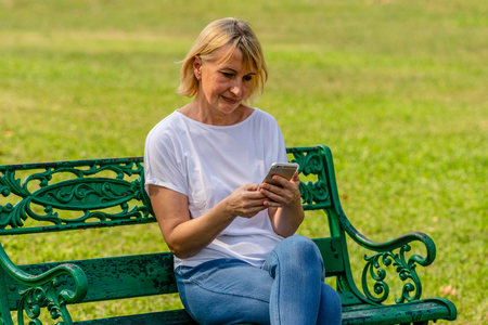 Elderly woman sitting on bench and using her mobile phone to text message in Public parkの写真素材