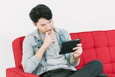 Asian Young man in denim jacket playing tablet sitting on Red sofa