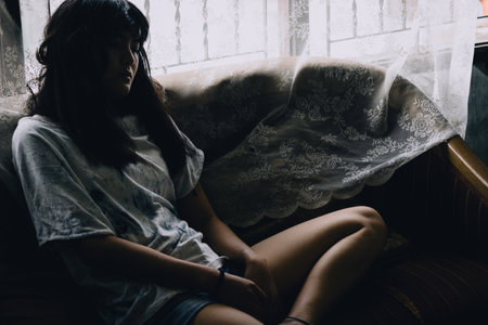 Asian girl sitting with absent mind on the sofa by the window with serious in Vintage cool toneの写真素材