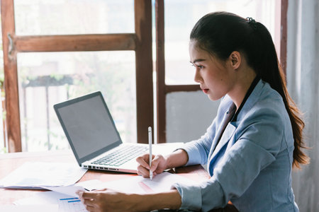 A long-haired Asian business woman is sitting at a desk with a notebook in front of her and hold a white pen and looking at the document plenty of documents on her deskの写真素材