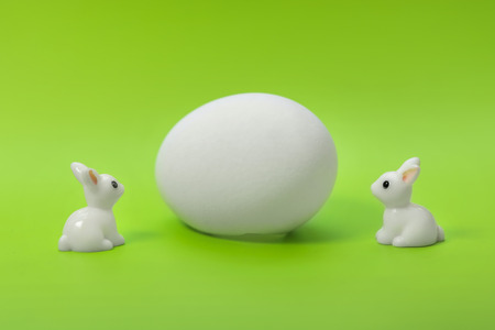 Two little white toy rabbits near the big white egg on a green backgroundの写真素材