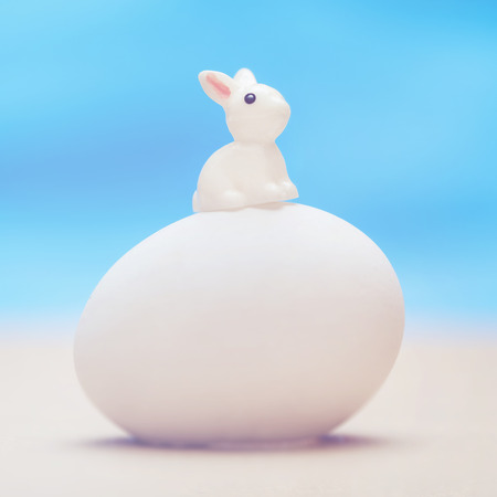 Little white toy rabbit sitting on a big white egg on a blue backgroundの写真素材