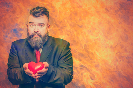 Scared adult bearded man in a suit holding a red heart in flames