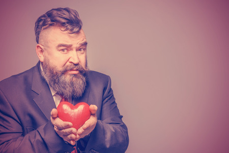 Scared adult bearded man in a suit holding a red heart. Tonedの写真素材