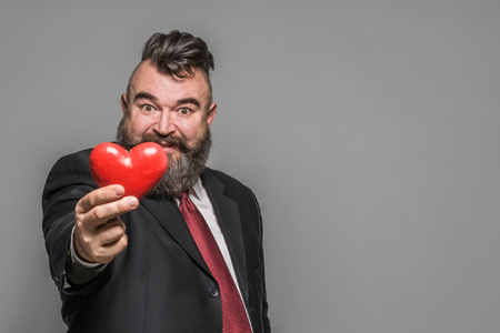 Adult bearded man in a black jacket and red tie holding red heartの写真素材