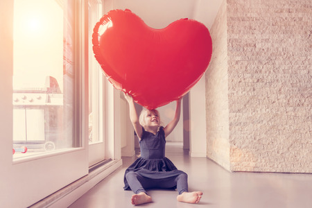 Little blonde girl in a blue dress holding a big red balloon in the shape of a heart  in the room near the windowの写真素材
