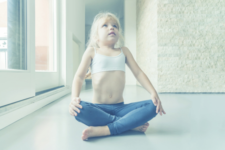 Little blond girl in white top and blue leggings doing a yoga exercise in the room near the windowの写真素材