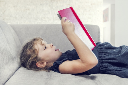 Little blonde girl is reading a red book on the couchの写真素材