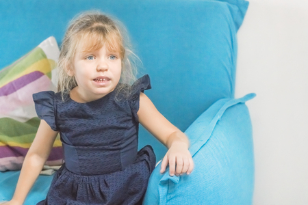 Little blonde girl in a blue dress sitting on the couch with a sad faceの写真素材