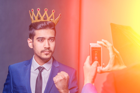 Young attractive man in a blue suit with a crown on his head photographed on phone in the sunlightの写真素材