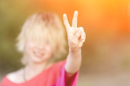 Girl blonde teenager in red blouse showing hand gesture victory in the sunlightの写真素材