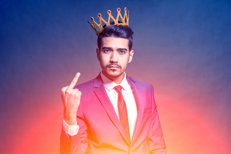 Young attractive man in a blue suit with a crown on his head showing middle finger in the bright...