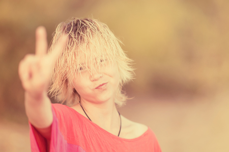 Girl blonde teenager in red blouse showing hand gesture victory in the sunlightの写真素材