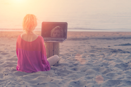 Girl blonde teenager in red blouse sitting in front of a laptop on the beach at sunsetの写真素材