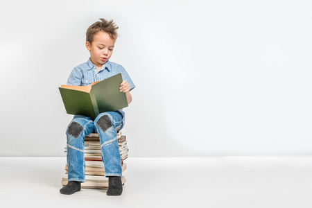 Cute boy in a blue shirt with a book sitting on a pile of booksの写真素材